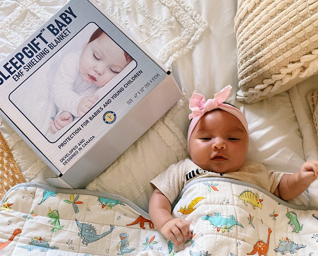A baby being wrapped by a SleepGift Baby EMF Protection Blanket as she lies on her bed, next to the packaging of the blanket.