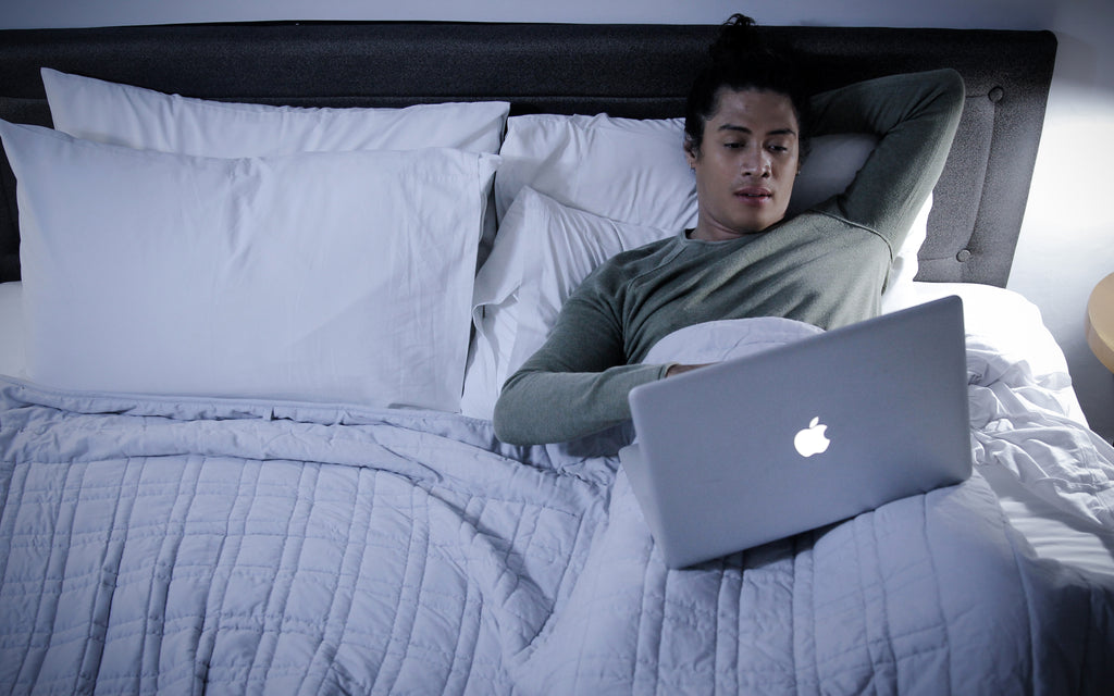 A man relaxing in bed under a SleepGift Weighted Blanket to protect himself from EMF radiation while using his laptop before bedtime. 